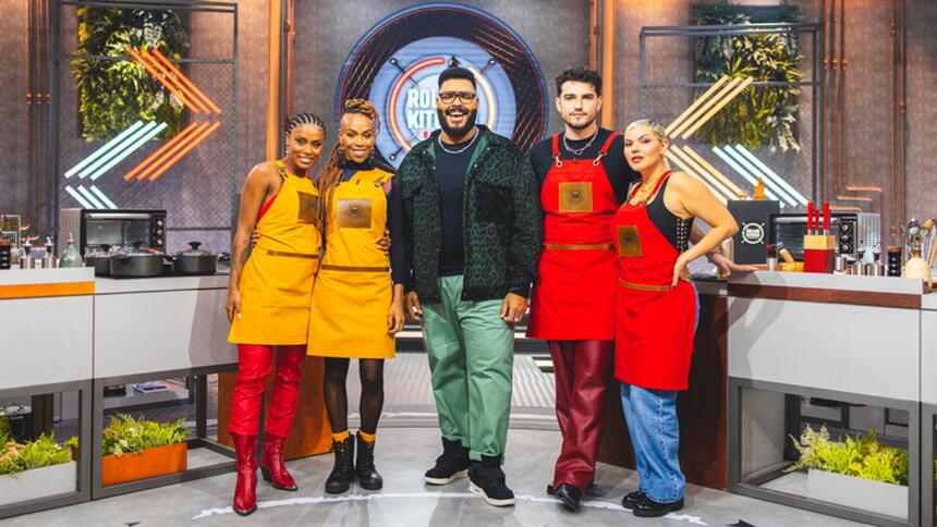 Rebecca sorrindo de calça e bota vermelha; Karol Conka com cabelo trançado, calça e bota preta; Paulo Vieira sorrindo, jaqueta e camisa preta, calça verde; Jão de camisa preta, calça vermelha e sapato preto, sorrindo; Duda Beat de camisa regata prta, calça jeans; Convidados estão usando avental vermelho com a logo do programa estampada