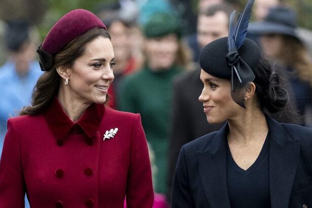 Kate Middleton de boina e sobretudo magenta ao lado de Meghan Makle de boina e sobretudo preto