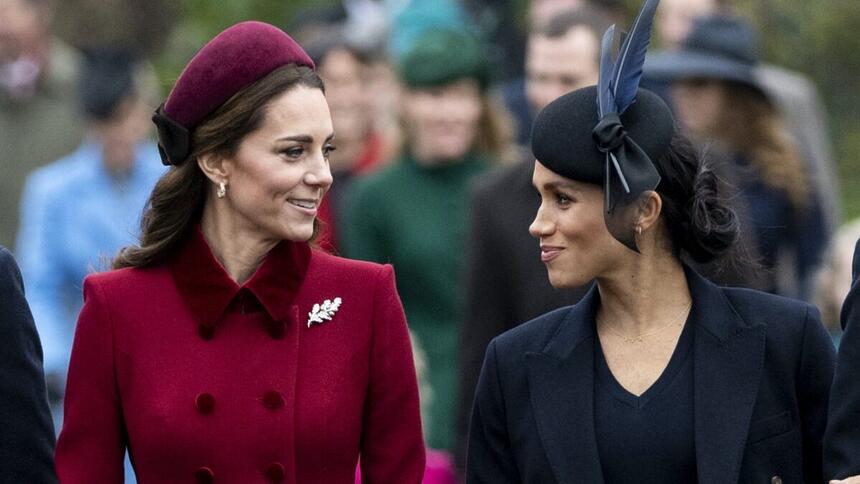 Kate Middleton de boina e sobretudo magenta ao lado de Meghan Makle de boina e sobretudo preto