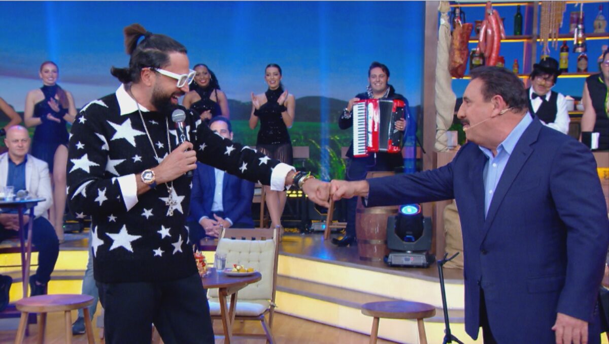 Latino com camisa manga longa preta com estampas de estrela branca dando soco na mão de Ratinho durante gravação do Boteco do Ratinho