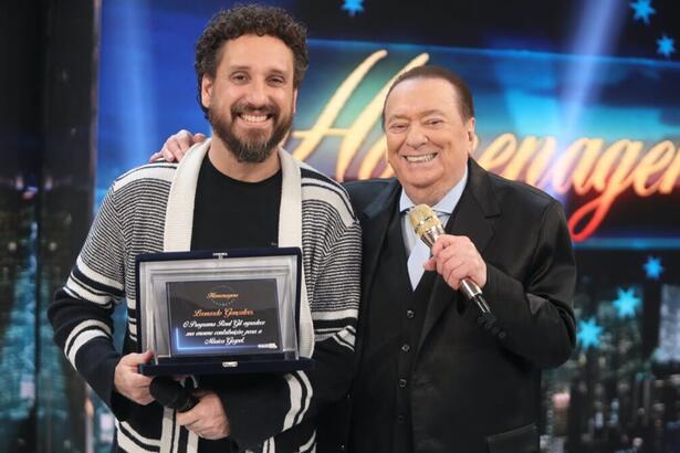 Leonardo Gonçalves sorrindo, de cardigã cinza e branco, com camisa preta, segurando uma placa de homenagem durante gravação do Programa do Raul Gil, do SBT