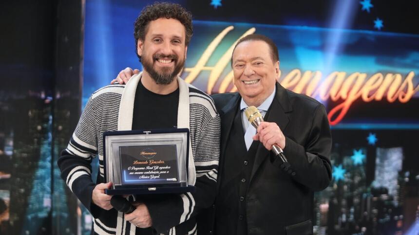 Leonardo Gonçalves sorrindo, de cardigã cinza e branco, com camisa preta, segurando uma placa de homenagem durante gravação do Programa do Raul Gil, do SBT