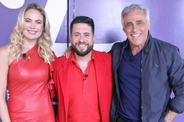 Oscar Magrini de blazer preto e camisa social azul, sorrindo; Thaís Pacholek de vestido vermelho, sorrindo ao lado de Mauricio Meirelles durante gravação do Foi Mau, da RedeTV!