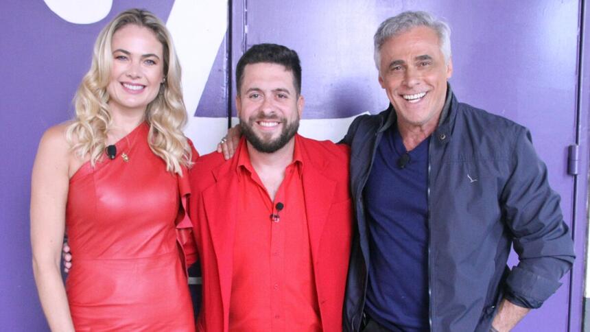 Oscar Magrini de blazer preto e camisa social azul, sorrindo; Thaís Pacholek de vestido vermelho, sorrindo ao lado de Mauricio Meirelles durante gravação do Foi Mau, da RedeTV!