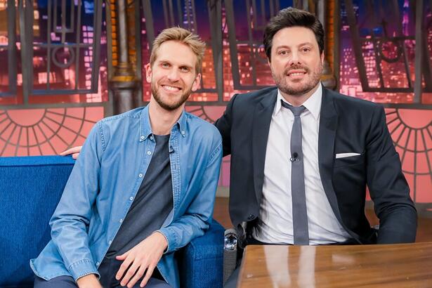 Paul Cabannes de jaqueta e calça jeans azul claro, camisa cinza, sorrindo, sentado, ao lado de Danilo Gentili durante gravação do The Noite, do SBT