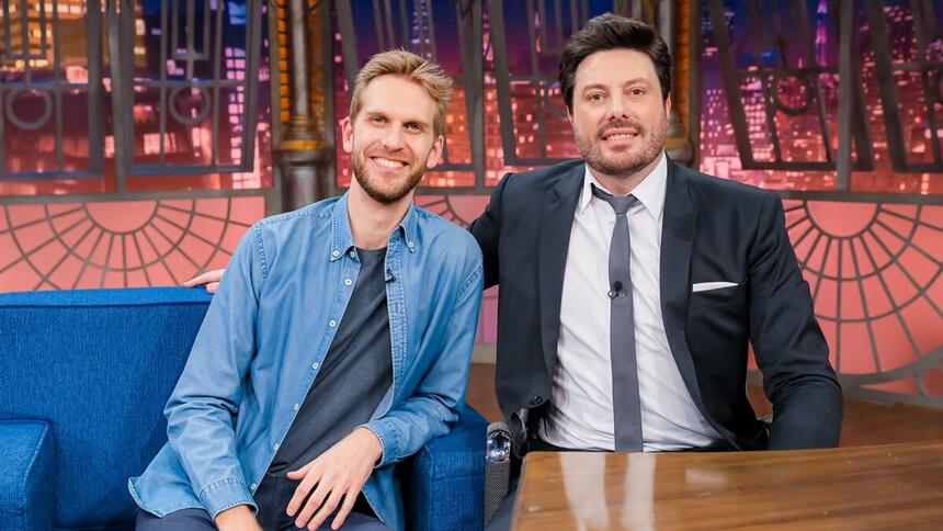 Paul Cabannes de jaqueta e calça jeans azul claro, camisa cinza, sorrindo, sentado, ao lado de Danilo Gentili durante gravação do The Noite, do SBT