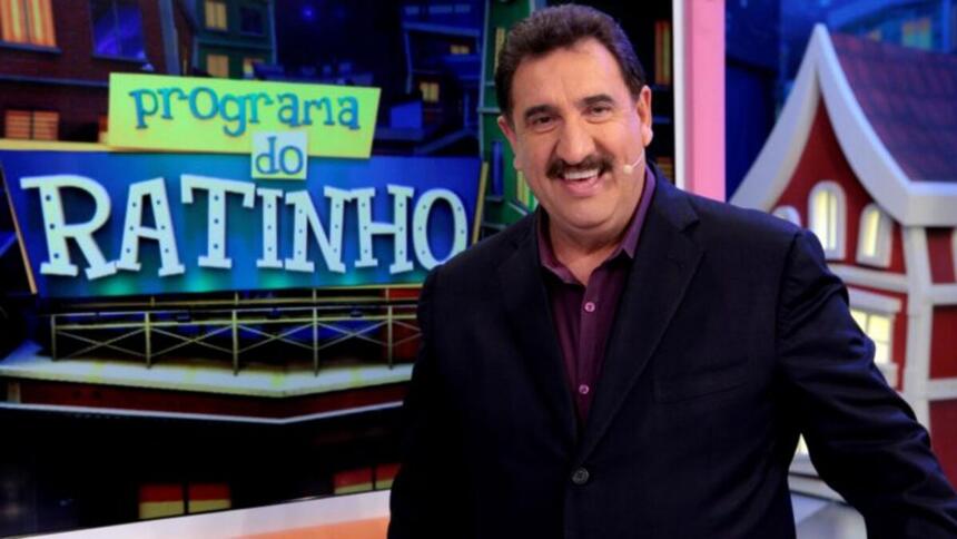 Ratinho de blazer preto e camisa social marrom, sorrindo, de perfil, microfone no rosto durante gravação de seu programa no SBT