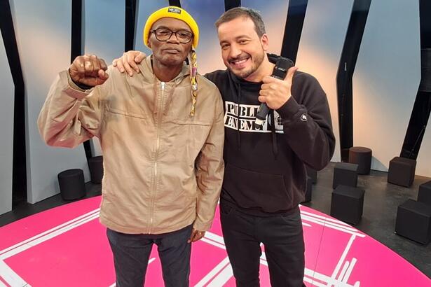 Rafael Cortez de camisa e calça preta sorrindo, dando um legal com a mão esquerda; Rappin Hood de camisa marrom, boina amarela, calça jeans preta durante gravação do Matéria Prima, da TV Cultura