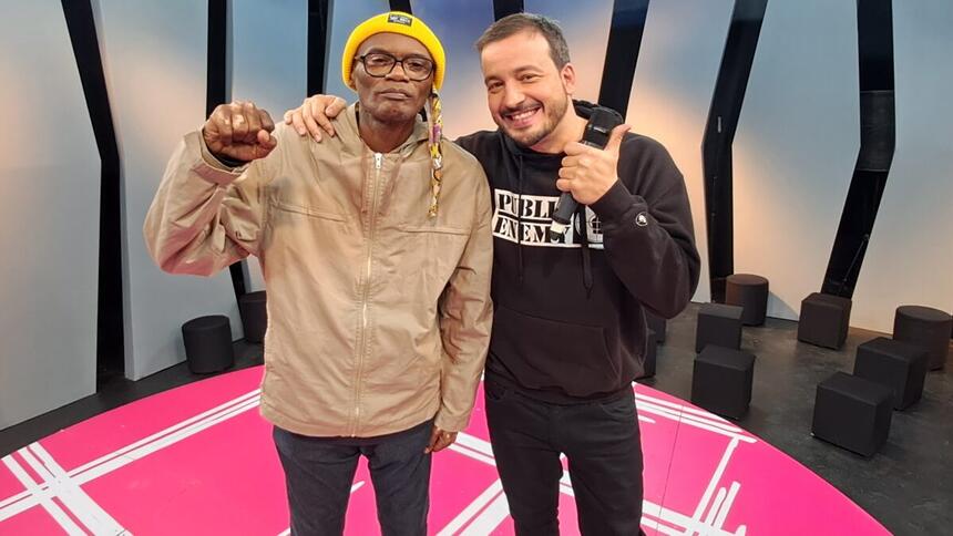 Rafael Cortez de camisa e calça preta sorrindo, dando um legal com a mão esquerda; Rappin Hood de camisa marrom, boina amarela, calça jeans preta durante gravação do Matéria Prima, da TV Cultura