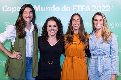 Renata Silveira, Natália Lara, Renata Mendonça e Ana Thais Matos juntos e abraçadas, em frente a um banner da Globo sobre a Copa