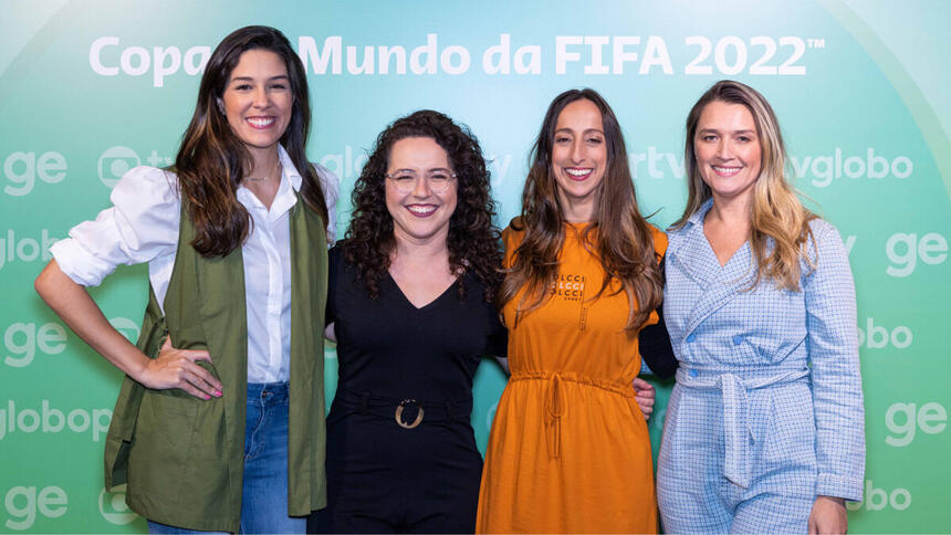 Renata Silveira, Natália Lara, Renata Mendonça e Ana Thais Matos juntos e abraçadas, em frente a um banner da Globo sobre a Copa