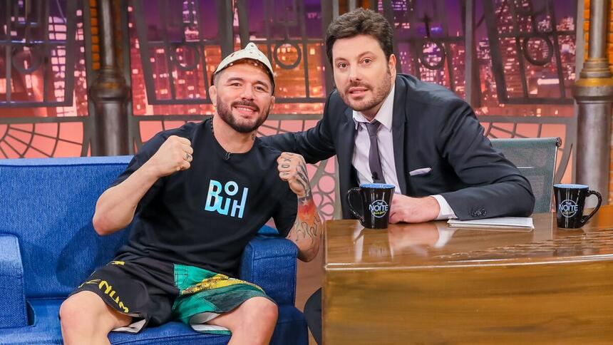 Rony Jason de short preto com estampa em verde e amarelo, camisa preta estampada na cor azul, fazendo um soco com as duas mãos e a máscara na cabeça; Danilo Gentili de palitó, gravata e calça preta e camisa social branca; ambos sentados durante gravação do The Noite do SBT