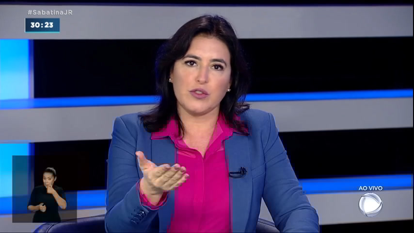 Simone Tebet de blazer azul, camisa social rosa, gesticulando durante entrevista na Record TV