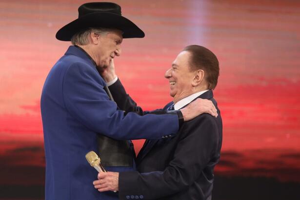 Sérgio Reis de lado, usando blazer azul escuro, chapéu preto com a mão no ombro de Raul Gil durante gravação do Programa Raul Gil, do SBT