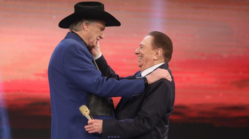 Sérgio Reis de lado, usando blazer azul escuro, chapéu preto com a mão no ombro de Raul Gil durante gravação do Programa Raul Gil, do SBT