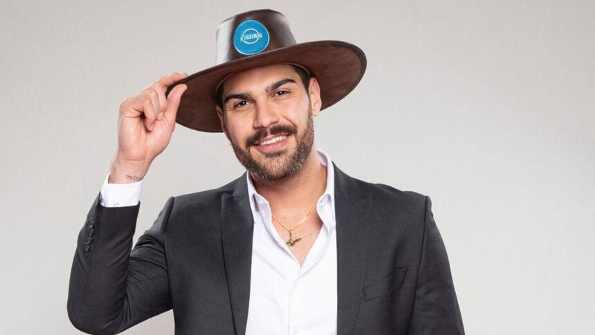 Shayan de blazer preto, camisa social branca, sorrindo, segurando seu chapéu de fazendeiro durante ensaio fotográfico de A Fazenda 14