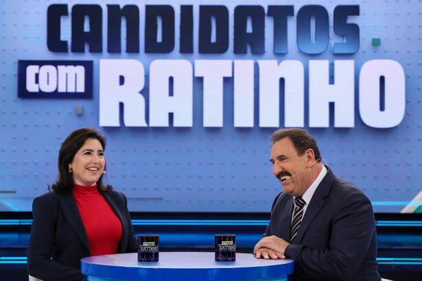 Simone Tebet de jaqueta preta, camisa vermelha, sorrindo, sentada em frente a Ratinho durante gravação do Candidatos com Ratinho, do SBT