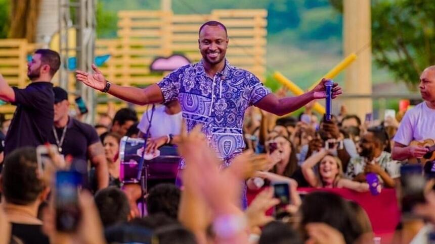 Thiaguinho sorrindo, de camisa roxa estampada, com braços abertos durante apresentação do Tardezinha