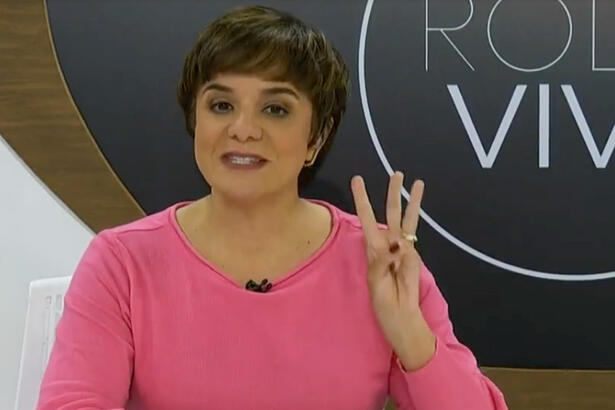 Vera Magalhães de camisa manga longa rosa, fazendo o numero 3 com a mão esquerda, durante gravação no estúdio do Roda viva, da TV Cultura