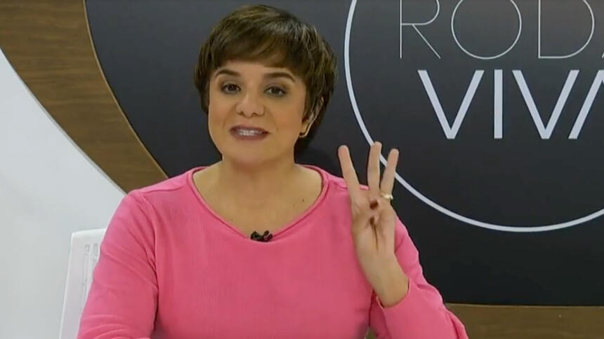 Vera Magalhães de camisa manga longa rosa, fazendo o numero 3 com a mão esquerda, durante gravação no estúdio do Roda viva, da TV Cultura