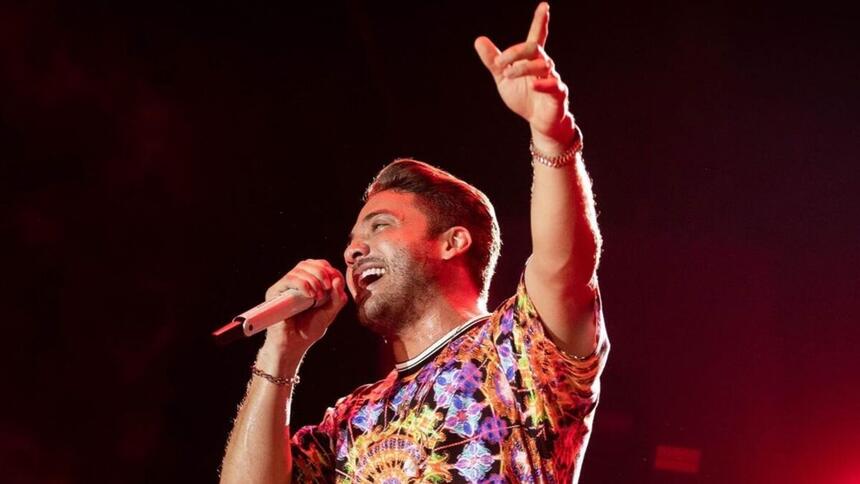 Wesley Safadão com camisa estampada colorida com o braço esquerdo levantado, apontando pra cima, segurando o microfone com a mão esquerda