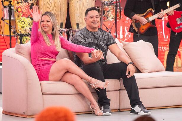 Xanddy de camisa cinza, calça preta, mão esquerda no joelho e mão direita dada a Carla Perez; Carla Perez de vestido rosa, sorrindo, acenando para a plateia durante gravação do Pipoca da Ivete, da TV Globo