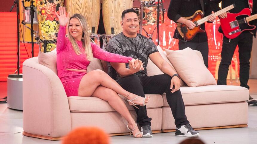 Xanddy de camisa cinza, calça preta, mão esquerda no joelho e mão direita dada a Carla Perez; Carla Perez de vestido rosa, sorrindo, acenando para a plateia durante gravação do Pipoca da Ivete, da TV Globo