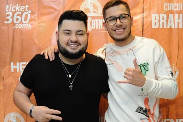 dom vittor e gustavo posando juntos, em frente a um banner nos bastidores de um show