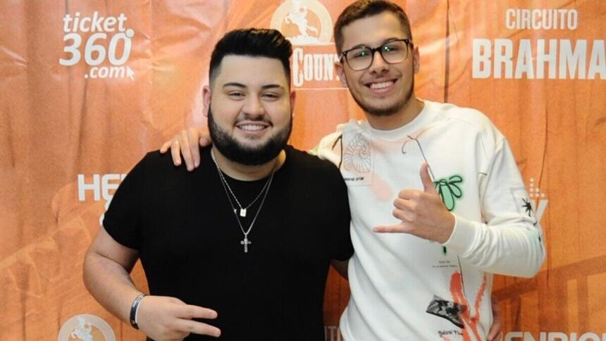 dom vittor e gustavo posando juntos, em frente a um banner nos bastidores de um show