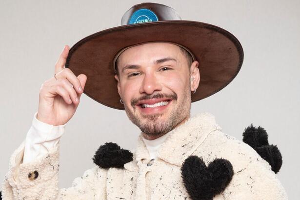 Alex com figurino inspirado em vaca, com jaqueta na cor de gelo e adereços de coração, com chapéu de fazendeiro e semblante sério