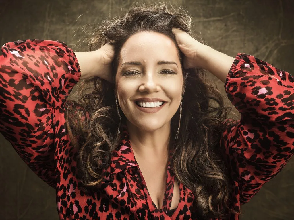 Ana Carolina sorrindo, com as mãos na cabeça, usando camisa estampada vermelha com preto