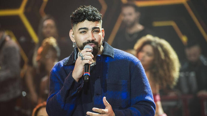 Dilsinho de camisa social azul e preta, segurando o microfone preto com a mão direita durante performance no Faustão da Band