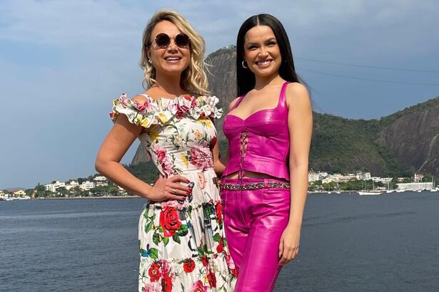 Eliana de óculos, vestido florido, com a mão na cintura ao lado de Juliette de top e calça rosa, sorrindo, durante entrevista