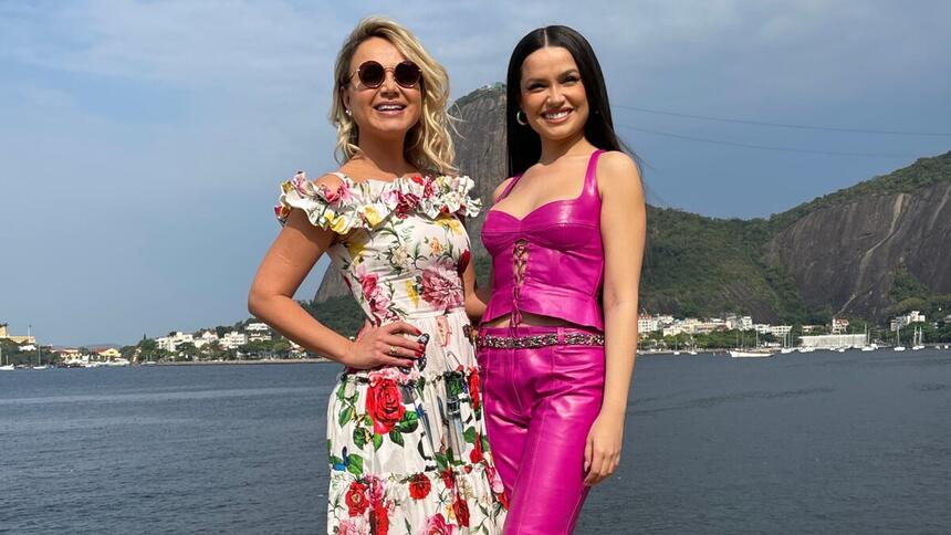 Eliana de óculos, vestido florido, com a mão na cintura ao lado de Juliette de top e calça rosa, sorrindo, durante entrevista