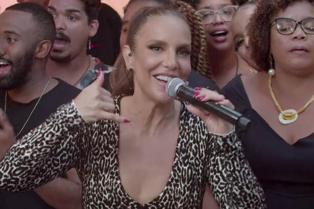 Ivete Sangalo de macacão animal-print fazendo um telefone com a mão direita segurando um microfone com a mão esquerda durante gravação do Macaco Sessions