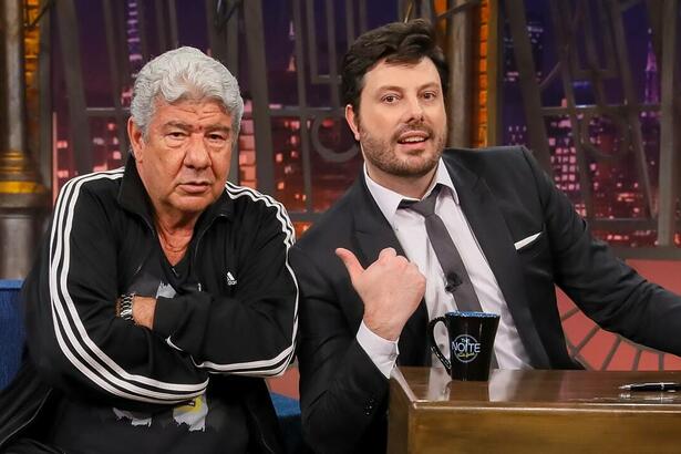 Joel Santana de camisa e calça preta, semblante sério, braços cruzados, ao lado de Danilo Gentili durante gravação do The Noite, do SBT