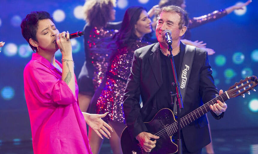 Kell Smith de rosa e Amado Batista de blazer e camisa pretos segurando uma guitarra, cantando durante gravação do Faustão na Band