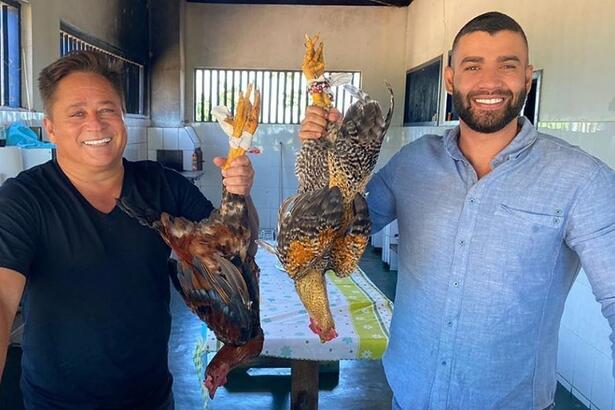 Leonardo sorrindo, de camisa preta segurando uma galinha ao lado de Gusttavo Lima