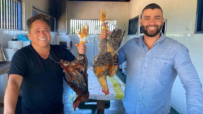 Leonardo sorrindo, de camisa preta segurando uma galinha ao lado de Gusttavo Lima