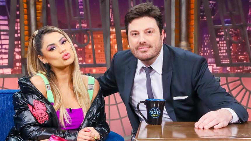 Lexa de look roxo e verde, fazendo bico com a boca, cabelo solto, sentada ao lado de Danilo Gentili durante gravação do The Noite