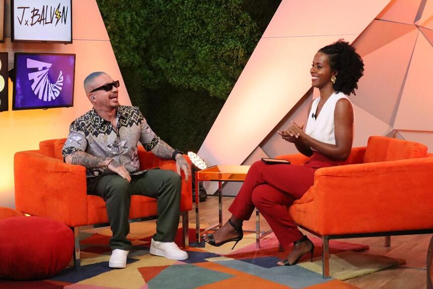 Maju de camisa branca e calça vermelha, sentada, sorrindo em frente a J Balvin durante gravação de entrevista para o Fantástico, da TV Globo