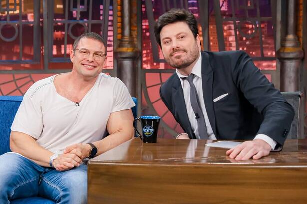 Paulo Muzy de camisa branca, calça jeans azul, sorrindo, de oculos de grau ao lado de Danilo Gentili durante gravação do The Noite, do SBT