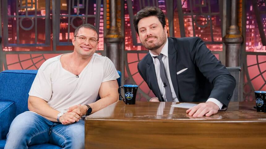 Paulo Muzy de camisa branca, calça jeans azul, sorrindo, de oculos de grau ao lado de Danilo Gentili durante gravação do The Noite, do SBT