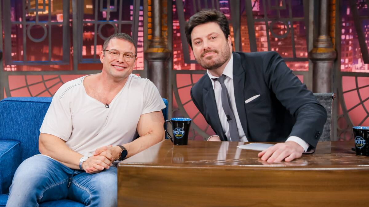 Paulo Muzy de camisa branca, calça jeans azul, sorrindo, de oculos de grau ao lado de Danilo Gentili durante gravação do The Noite, do SBT