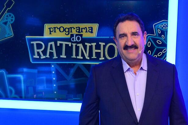 Ratinho de blazer azul escuro e camisa social azul clara, sorrindo ao lado da logo do seu programa durante gravação