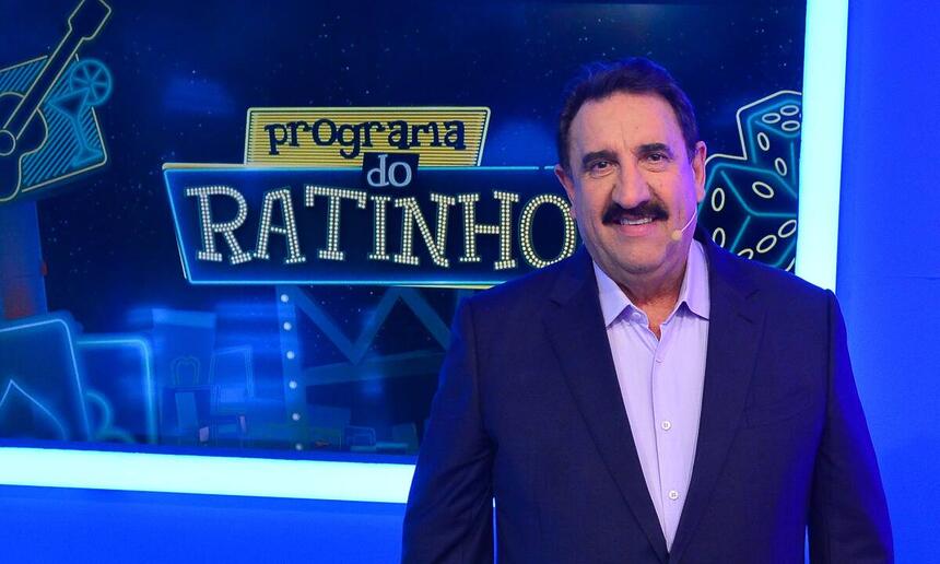 Ratinho de blazer azul escuro e camisa social azul clara, sorrindo ao lado da logo do seu programa durante gravação