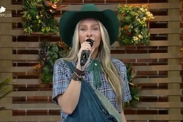 Adriane Galisteu com microfone na mão com chapéu e semblante sério.