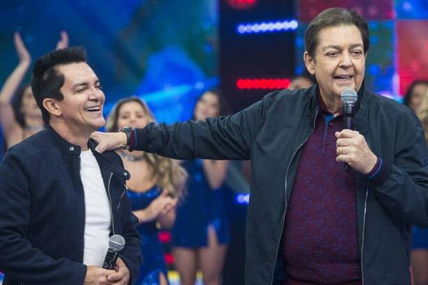 Regis Danese sorrindo, de camisa branca e preta, ao lado de Faustão durante gravação de programa