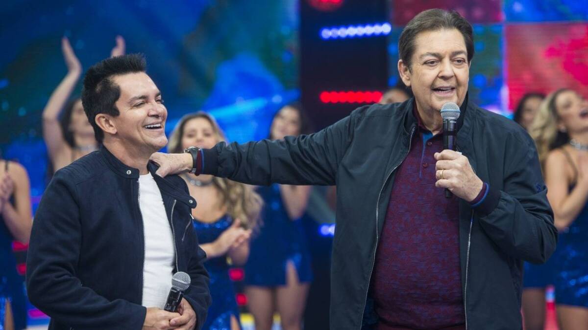 Regis Danese sorrindo, de camisa branca e preta, ao lado de Faustão durante gravação de programa