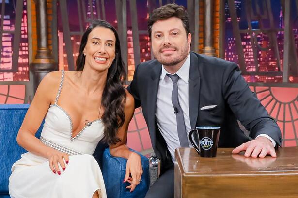 Sheilla Castro de vestido branco, sorrindo, com a mão no braço do sofá azul ao lado de Danilo Gentili durante gravação do The Noite, do SBT